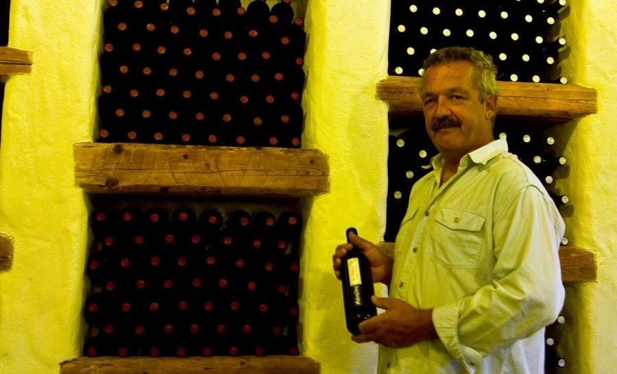 man keeping a bottle of wine front 'Manalis Winery' cellar with wine bottles on top of each other storaged in the stone wall